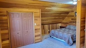 A bedroom with wooden walls and a sloped ceiling, featuring a double door, a bed with patterned bedding, and a small nightstand.