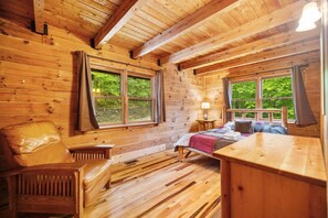 Ein gemütliches Blockhausszimmer mit einem Bett, einem Sessel, einem kleinen Tisch und zwei Fenstern mit Blick auf Grünflächen.