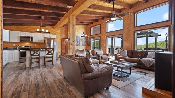 A spacious living area with a large brown leather sofa, a coffee table, and a TV. There is a kitchen area with white cabinets and a microwave. The room has wooden floors and large windows offering a view of the outdoors. - Nearly 360 degree Mountain View! Private setting, Big Hot Tub, Wi-Fi, Gas Grill (Campton)