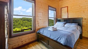 A wooden cabin bedroom with a large bed, two windows, and a view of a forest. - Nearly 360 degree Mountain View! Private setting, Big Hot Tub, Wi-Fi, Gas Grill (Campton)