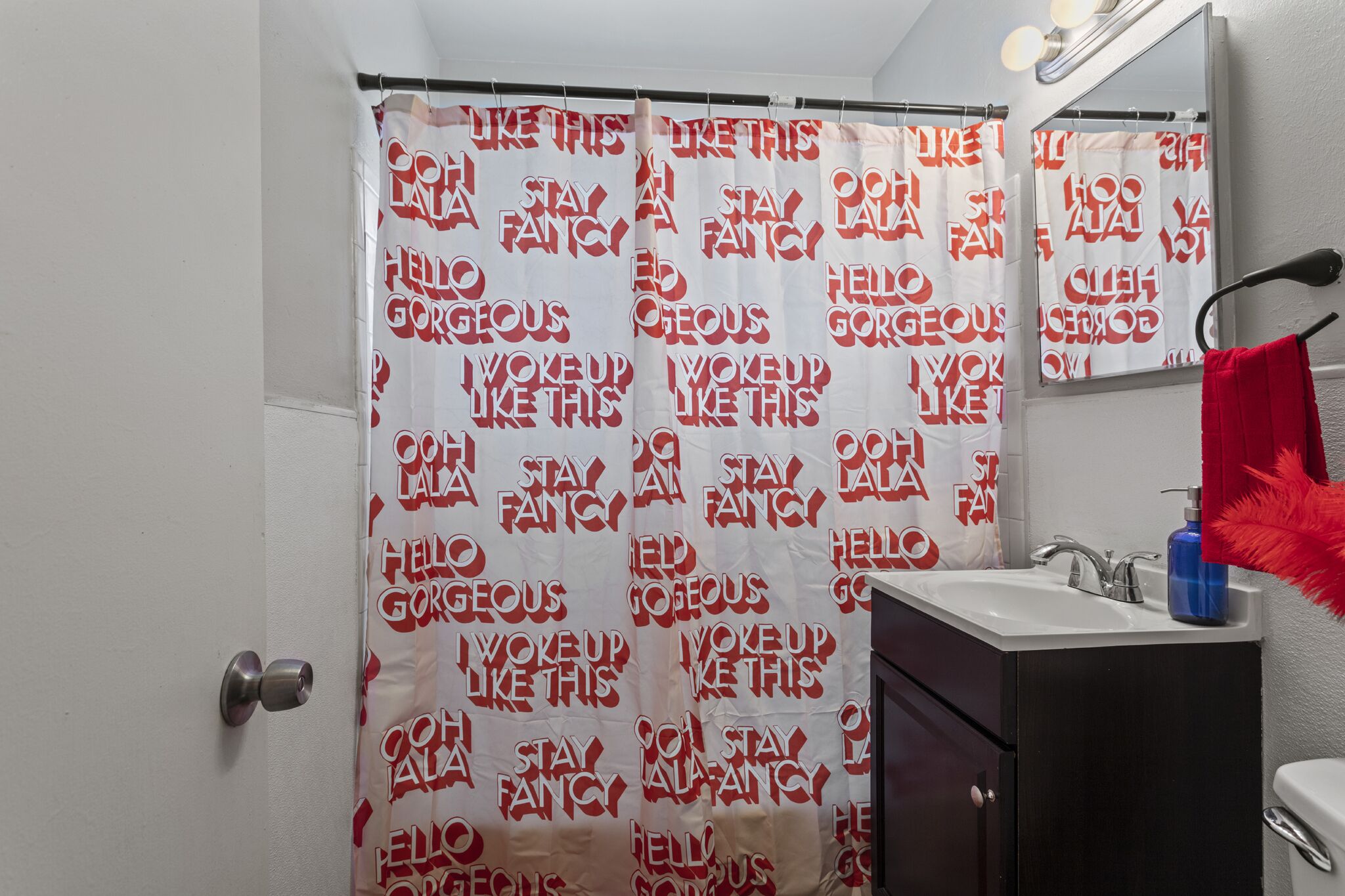 Een badkamer met douchegordijn met opvallende tekst, een wastafel met spiegel en een rode handdoek aan de muur.
