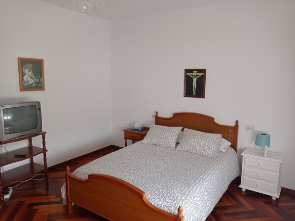 Un dormitorio con una cama de madera, una mesita de noche con lámpara, un televisor sobre un mueble de madera, y dos cuadros enmarcados en la pared.