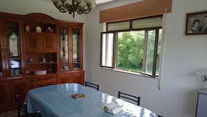Un coin repas avec une grande table recouverte d’une nappe bleue, entourée de chaises en bois. Un buffet en bois avec des portes en verre se trouve à gauche, et une fenêtre donnant sur la verdure se trouve à droite.