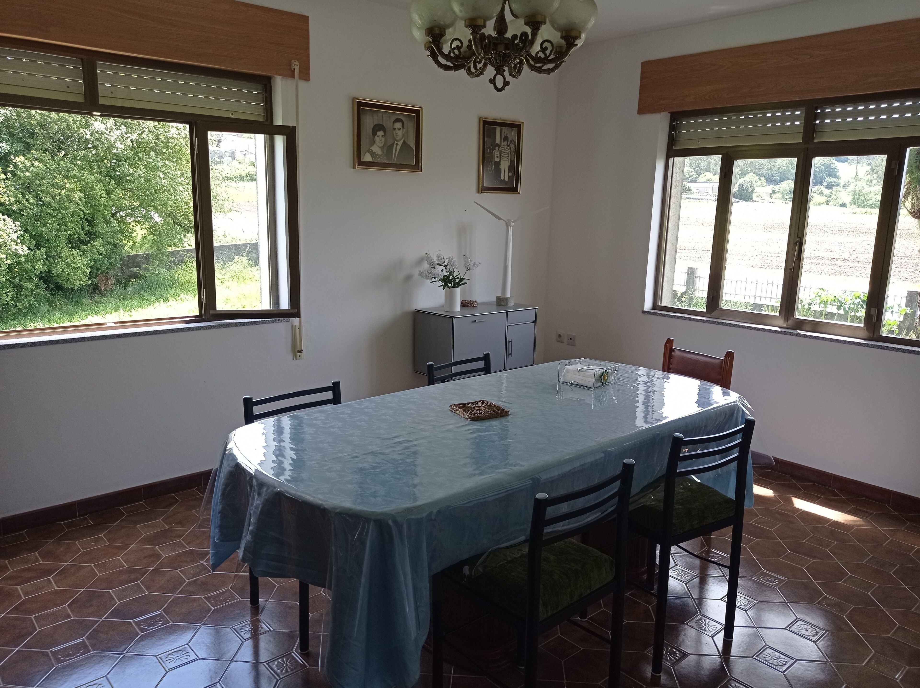 Ein Essbereich mit Tisch, Stühlen und einer Chandelier. Zwei Fenster mit Jalousien, und eine kleine Vitrine mit einer Topfpflanze.