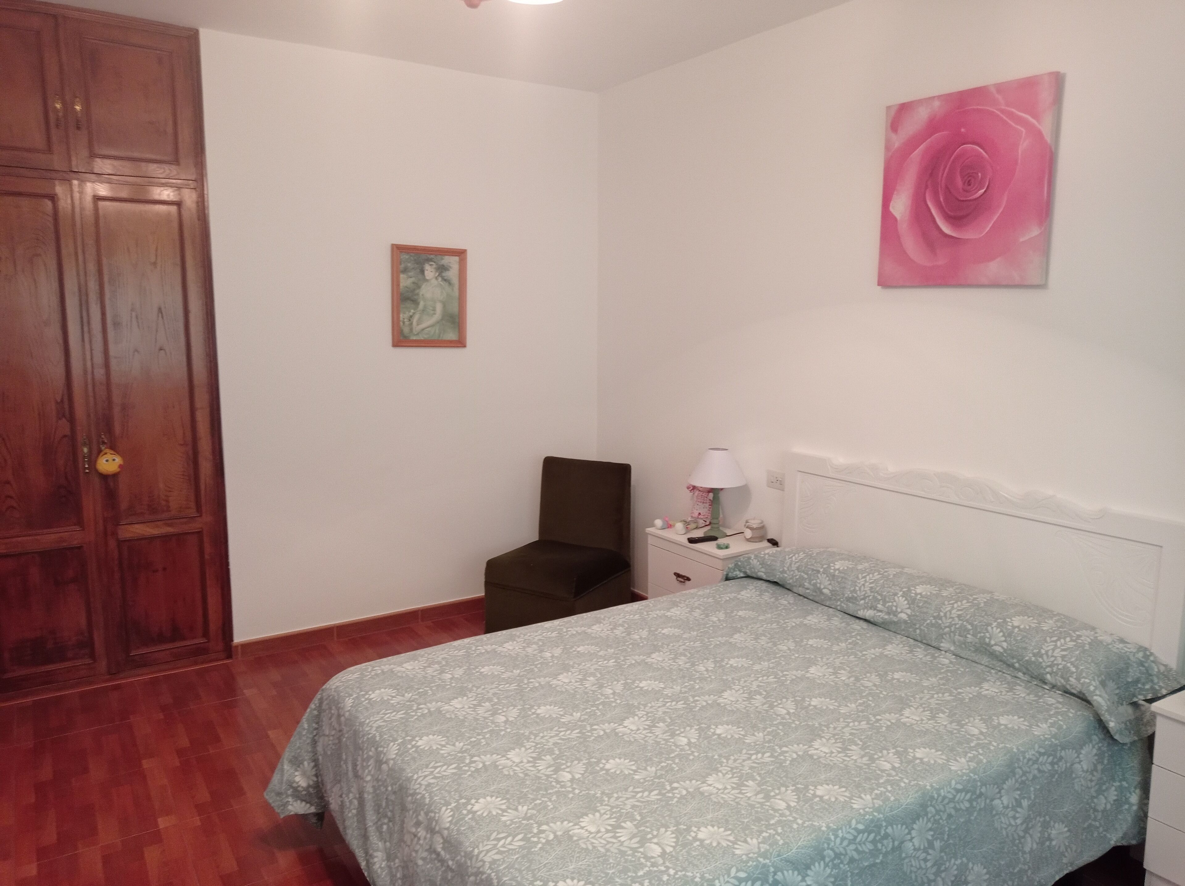 Een slaapkamer met een bed, nachtkastje, stoel, houten garderobe en een schilderij aan de muur.