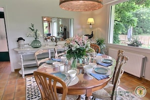 A dining area with a round table set for four, surrounded by wooden chairs. A vase with flowers is placed on the table, and a lamp is on the wall.
