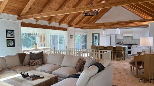 A spacious living area with a wooden ceiling, a large sofa, a coffee table, and a kitchen in the background. - Spacious house with pebble beach. Direct access to Snowmobile and ATV trails! (Beaver Cove)