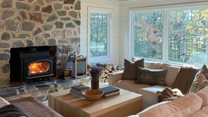 A cozy living room with a stone fireplace, wooden beams, and a large window offering a view of trees. - Spacious house with pebble beach. Direct access to Snowmobile and ATV trails! (Beaver Cove)