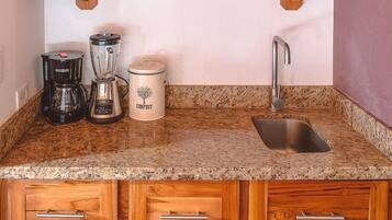 Eine Küche mit Granitarbeitsplatten, Holzausbau, einem Edelstahlspülbecken, einer Kaffeemaschine, einem Mixer, einer Komposttonne und einer Pendelleuchte.