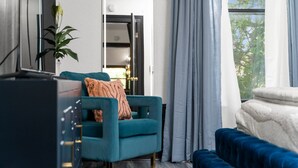 Una sala de estar moderna con un sillón azul verdoso, un sofá azul, un espejo grande y vistas a árboles a través de una ventana.