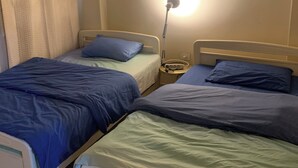 Een kamer met twee eenpersoonsbedden, elk met een blauwgroene spreidekker, een nachtkastje met een lamp en een airconditioner aan de muur.