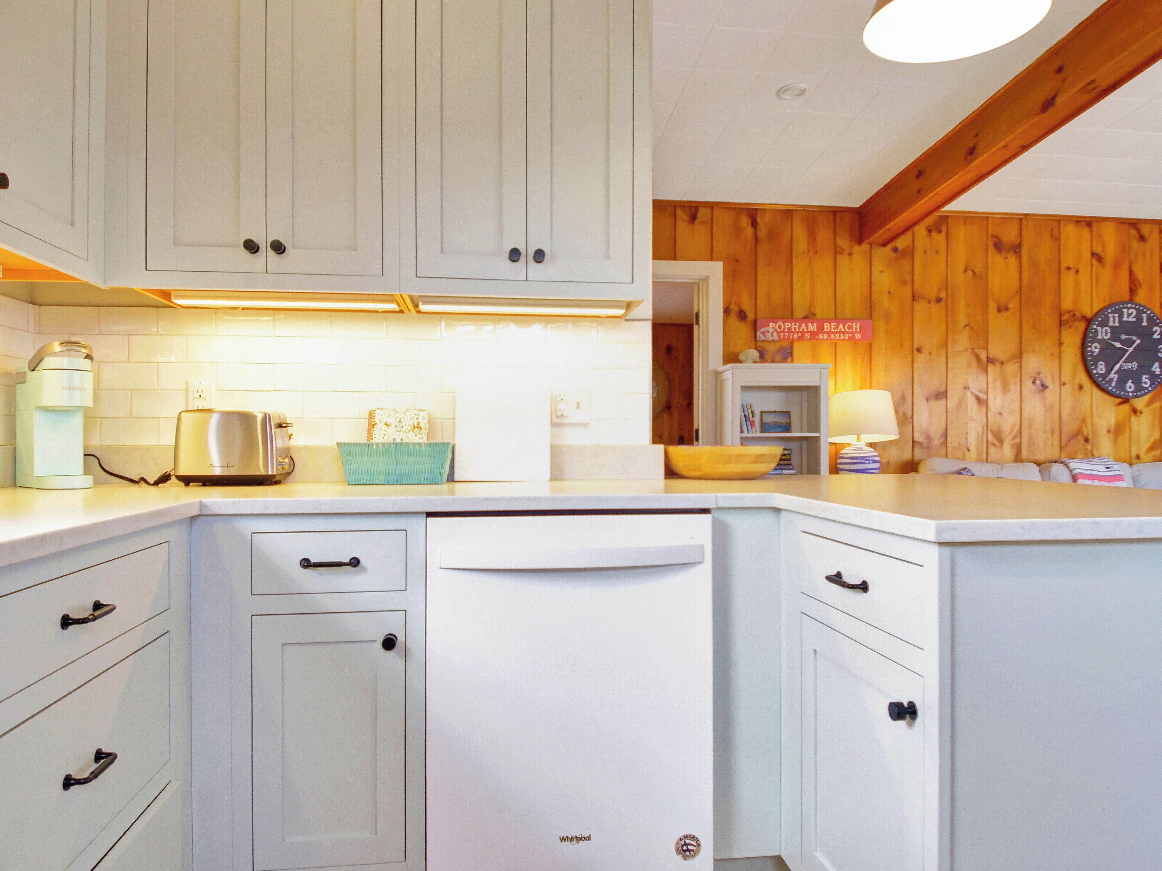 A kitchen with white cabinets, a dishwasher, a toaster, and a coffee maker. There is a wooden wall and a clock on the wall.
