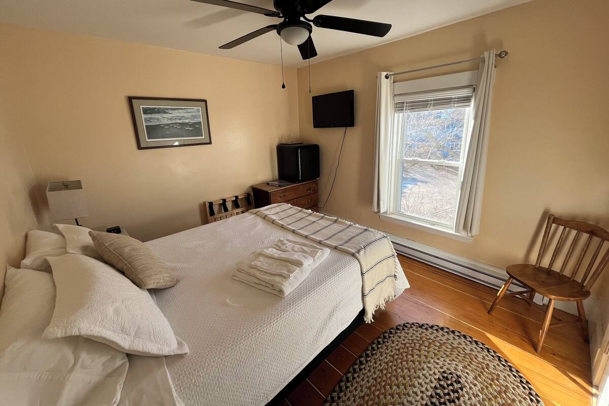 A hotel room with a bed, a ceiling fan, a TV, a window with curtains, and a wooden chair.