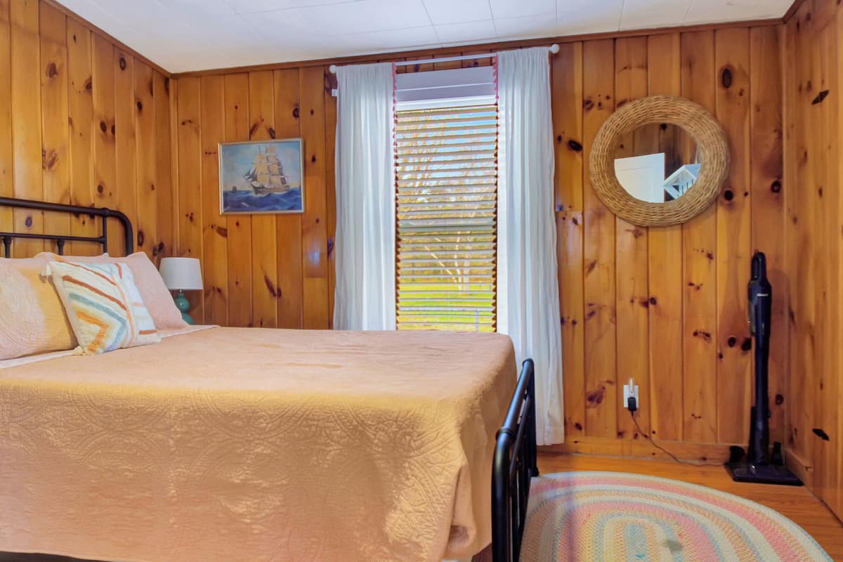 A wooden-paneled room with a bed, a painting, a window with blinds, a mirror, and a vacuum cleaner.