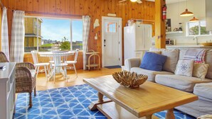 Un salon chaleureux avec un décor intérieur en bois, un coin repas avec des chaises blanches et une cuisine équipée d’un réfrigérateur et d’un évier.