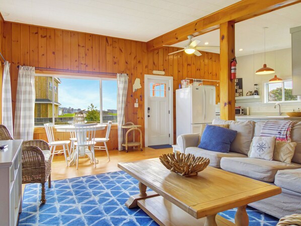 Una sala de estar acogedora con detalles de madera, un comedor con sillas blancas y una cocina equipada con refrigerador y pileta.