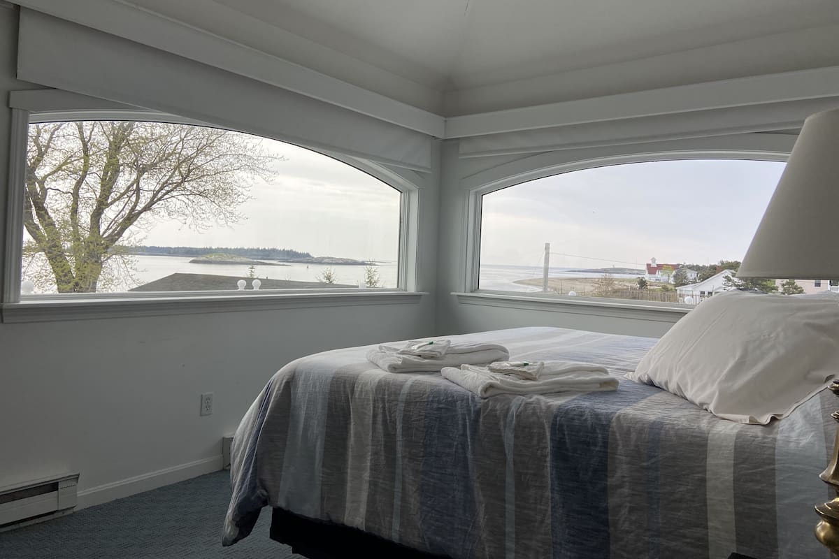 A bedroom with a large bed, two windows with a view of a body of water and a tree, and a ceiling fan.