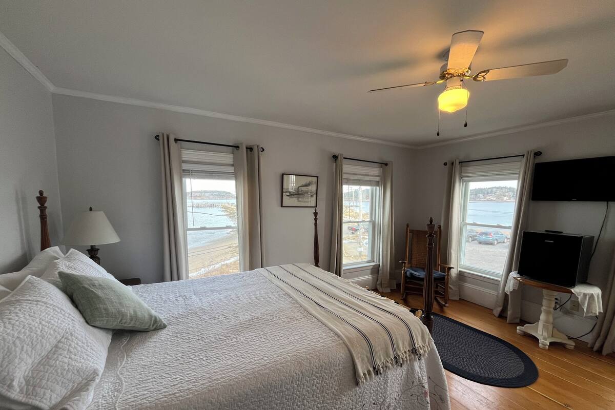 A hotel room with a bed, a ceiling fan, a TV, and a view of the sea.