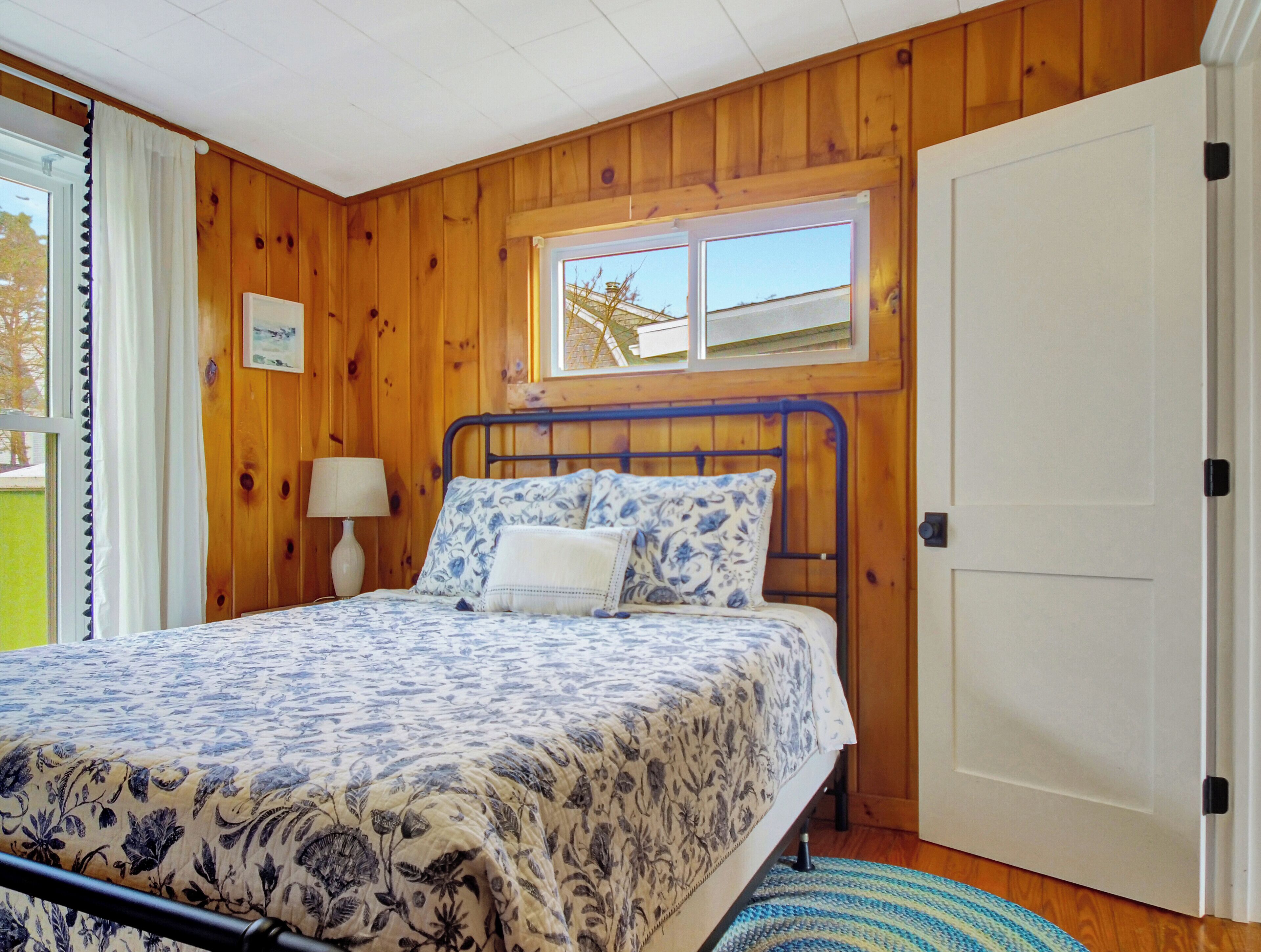 A bedroom with a wooden paneled wall, a bed with a floral bedspread, a window with a view, and a door.