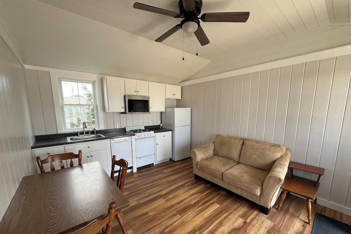 A compact kitchen with white cabinets, a stove, and a refrigerator. There is a wooden dining table with chairs and a beige sofa in a living area.