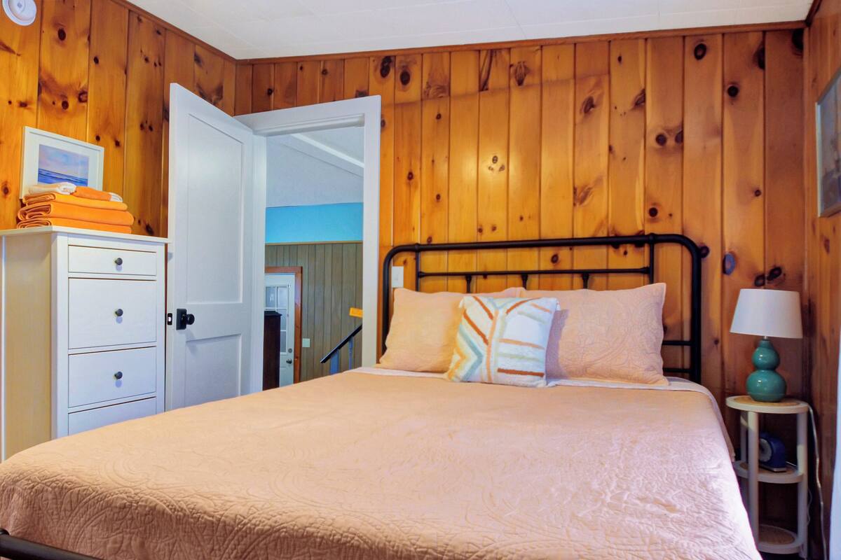 A bedroom with a bed, a nightstand with a lamp, a dresser, and a door leading to another room.