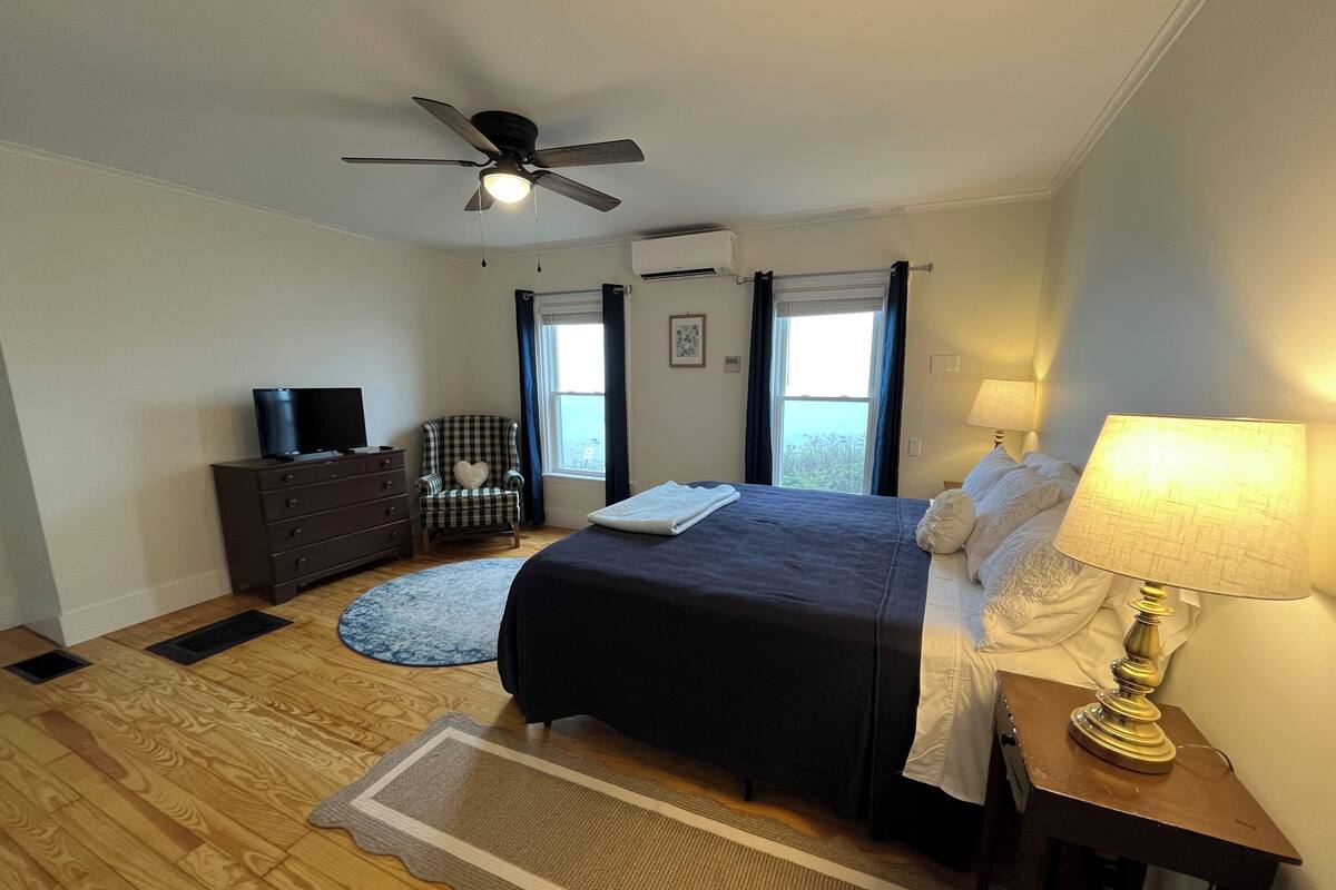 A bedroom with a bed, a television on a dresser, a chair with a heart pillow, a window with curtains, and a ceiling fan.