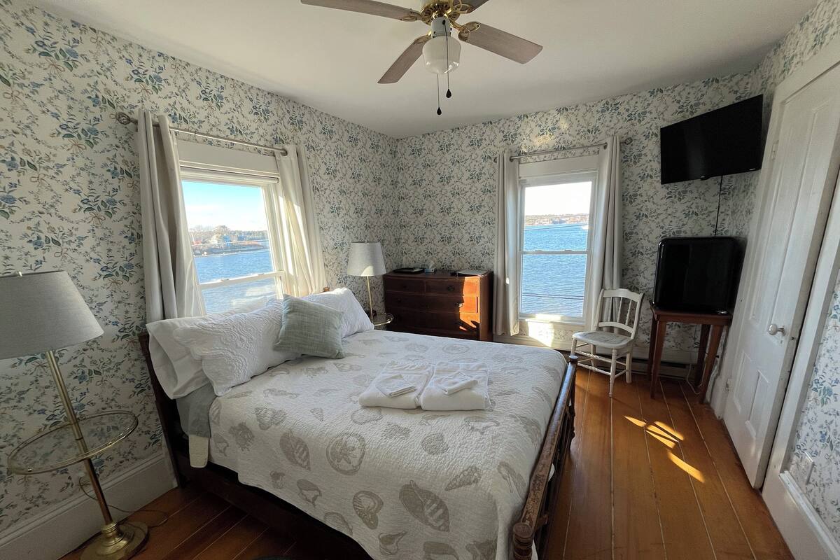 A bedroom with a bed, a desk, a chair, a TV, and a view of the water.