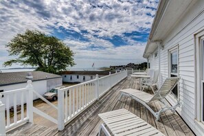 Una terraza de madera con barandas blancas y mobiliario exterior, con vistas a un cuerpo de agua y una costa lejana.