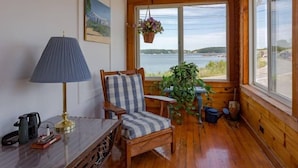 Une pièce chaleureuse dans un chalet avec des murs en bois, une vue sur un lac, un fauteuil avec des coussins à rayures, une table avec une lampe et une plante suspendue.