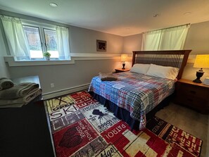 A bedroom with a bed, two bedside lamps, a window with curtains, a small bedside table, and a rug with printed patterns.