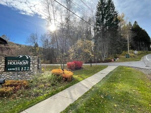 A welcome sign for Holimont with a stone wall, a paved pathway, and autumn foliage.