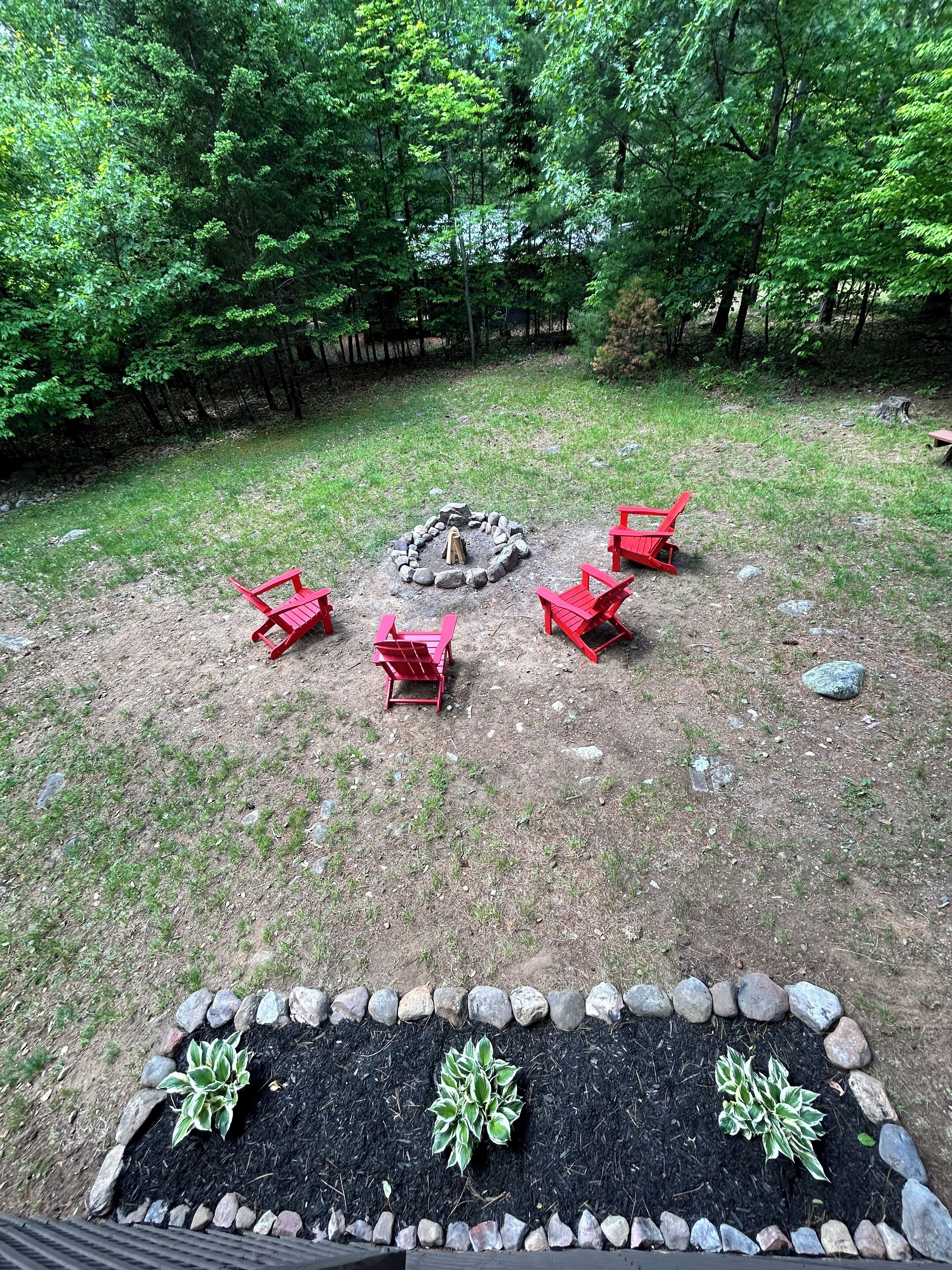 A backyard with a fire pit surrounded by red chairs, a stone border, and a garden bed with plants.