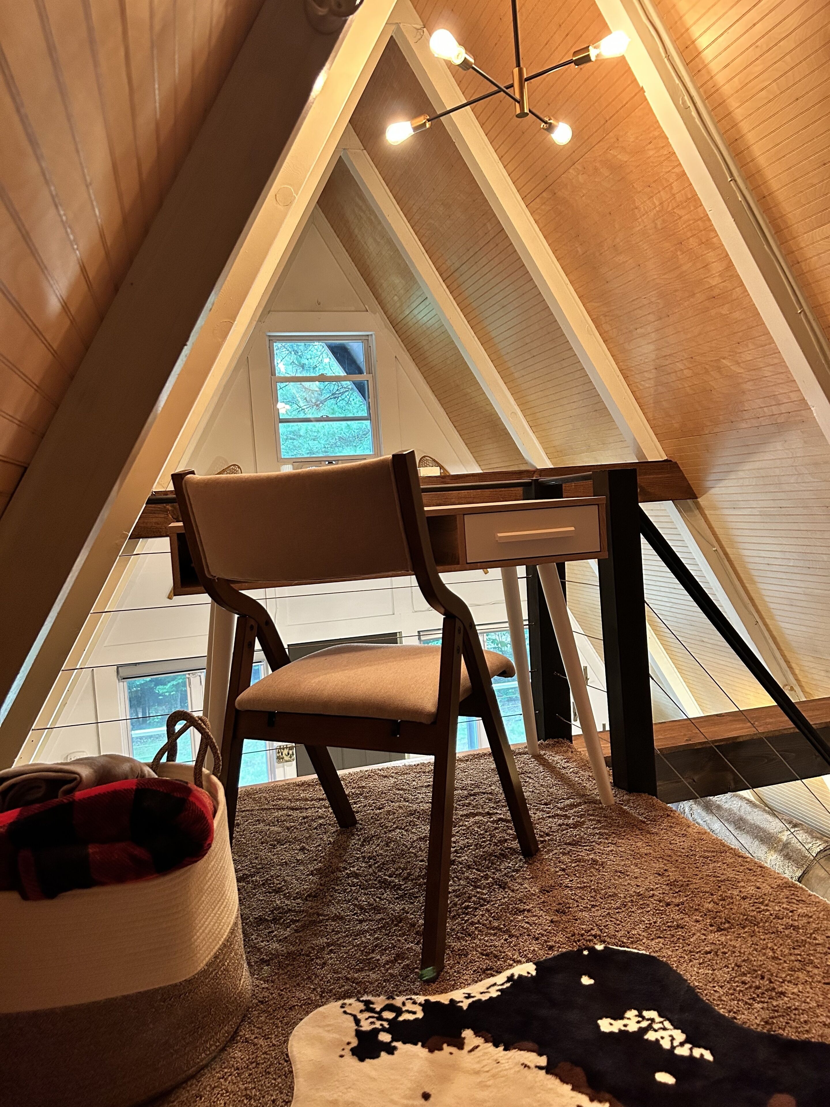 An attic room with a wooden ceiling, a chair, a table, and a window.