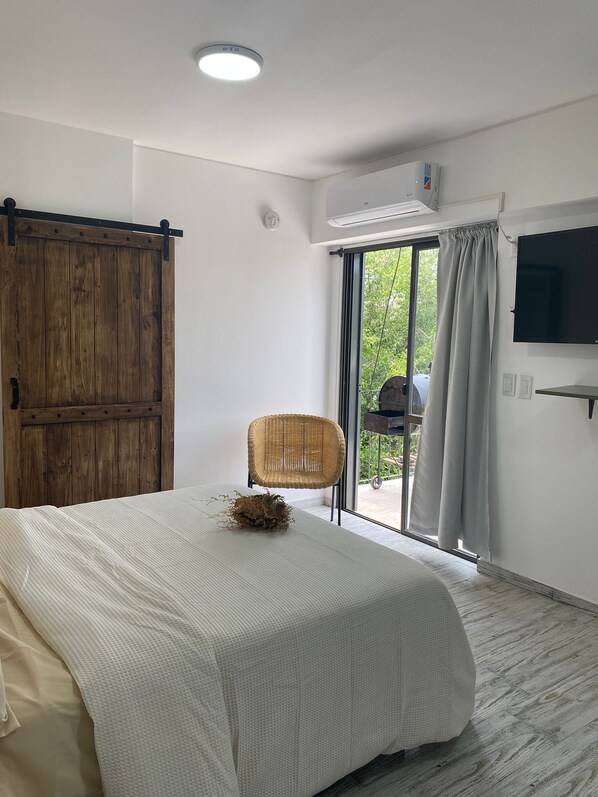 A bedroom with a bed, a wicker chair, a sliding door, a TV, and a window with curtains.