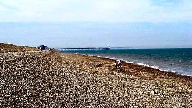 Une plage de galets, une jetée qui se dessine à l’horizon et un ciel dégagé.