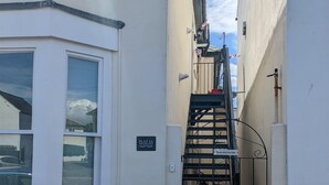 Une ruelle étroite avec un escalier menant à une entrée fermée par un portail. On aperçoit un pot de fleurs posé sur le sol.
