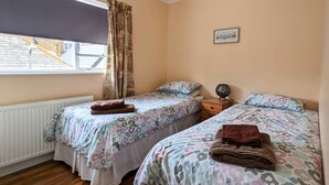 Une chambre avec deux lits, une fenêtre avec des rideaux, un radiateur et une petite table de chevet surmontée d’une lampe décorative.