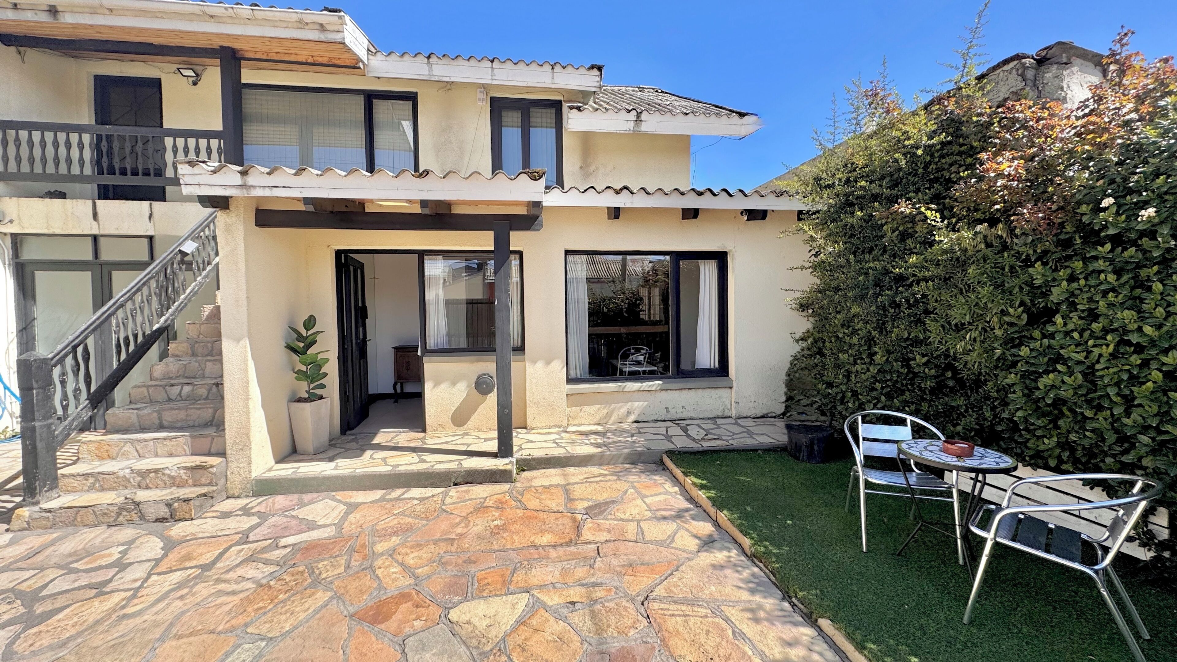 Una casa de dos pisos con un patio de piedra, un pequeño jardín y un área de comedor con mobiliario exterior.