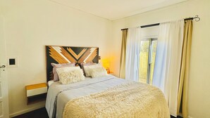 Un dormitorio con cama, cabecera de estampado geométrico, mesita de noche con lámpara y ventana con cortinas.