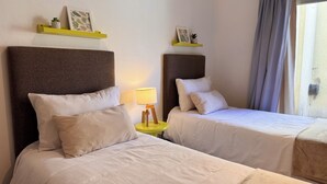 Habitación de hotel con dos camas, cada una con cabecero marrón y ropa de cama blanca. Hay mesitas de noche amarillas con lámparas y objetos decorativos.
