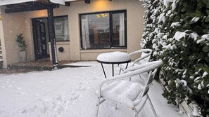 Een terras buiten, bedekt met sneeuw, met een tafel, stoelen, een bloempot en een gebouw met raam en deur.