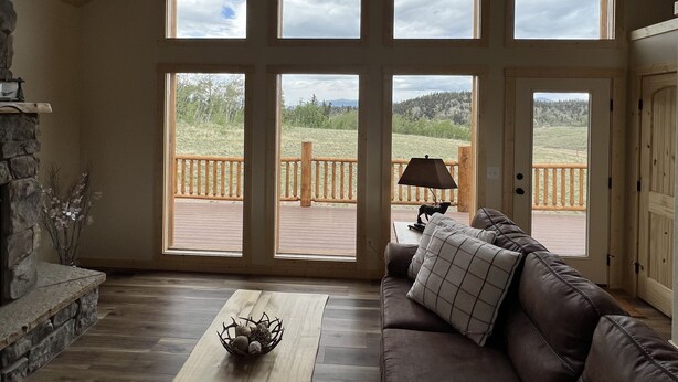A spacious living room with a stone fireplace, a wooden coffee table, and a leather sofa. Large windows offer views of a deck and surrounding landscape. - Brand New 4-bedroom cabin/stunning views/king bed, Hot Tub!!! (Jefferson)