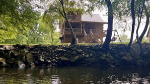 Een houten chalet met balkon, gelegen aan een waterpartij, omringd door bomen.