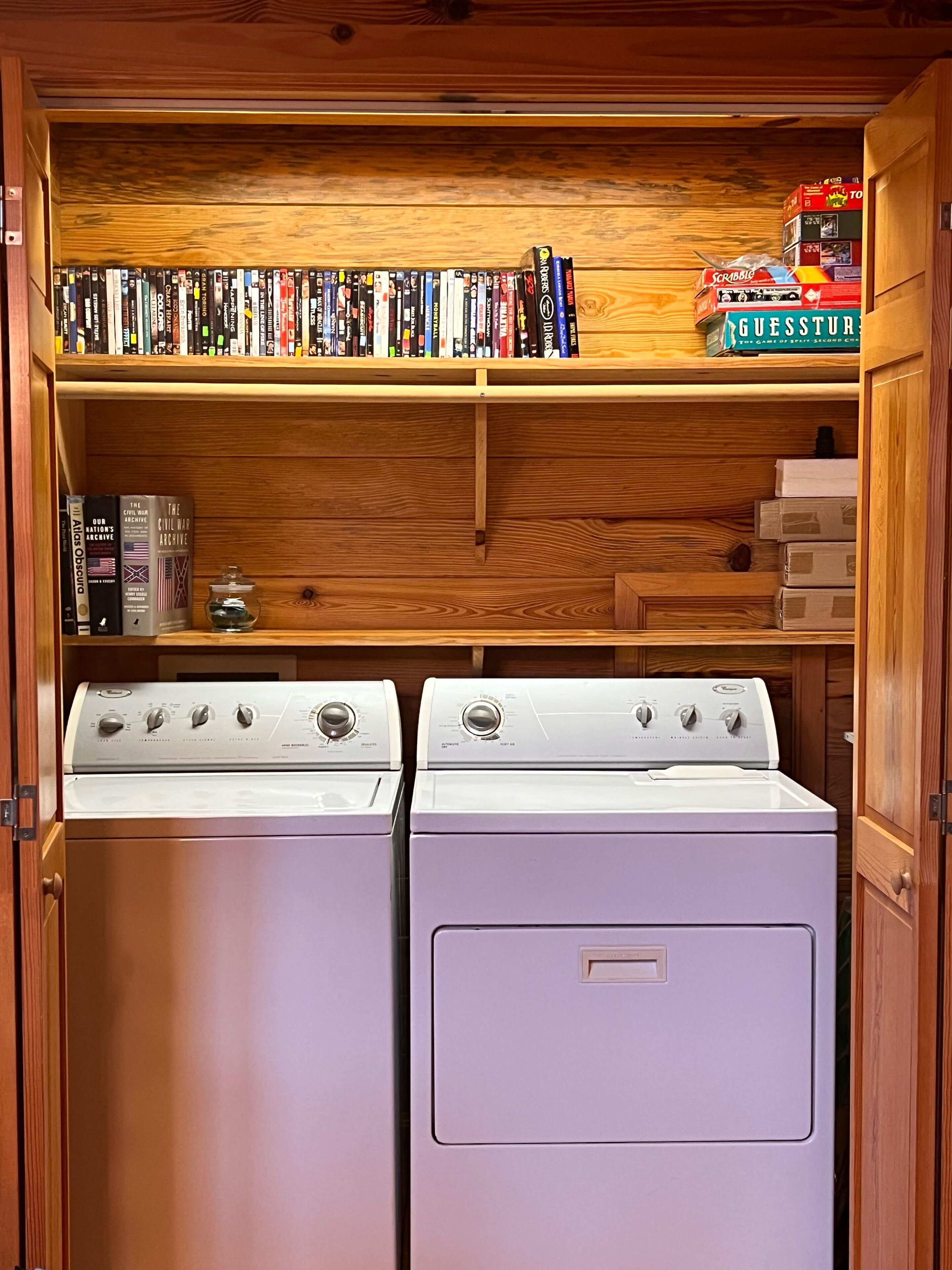 A laundry room with two stacked washers and dryers, wooden shelves above with books and a Scrabble box, and a small shelf with a jar and books.
