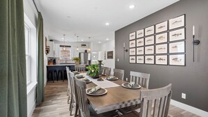 A dining area with a long wooden table set for six, surrounded by wooden chairs. A kitchen is visible in the background with modern appliances and a bar area. - Lakeview Home: Private Hot tub and Sauna (Branson)