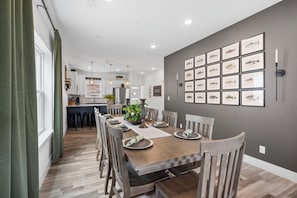 Een eetgedeelte met een lange houten tafel voor zes personen, omringd door houten stoelen. Achtergrond toont een keuken met moderne apparatuur en een bar.