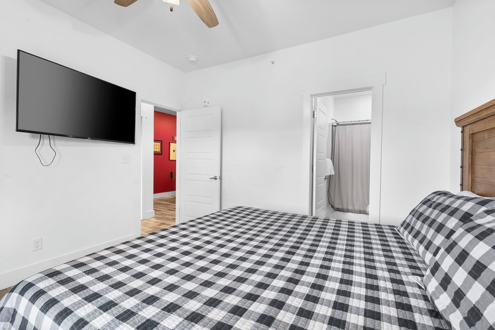 Een slaapkamer met een geruite sprei, een flatscreen televisie aan de muur en een houten plafondventilator.