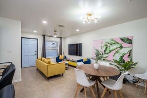 A modern living room with a yellow sofa, a blue sofa, a dining table with chairs, and a large plant. - Art Deco 3 BR Near Fort Bliss (El Paso)