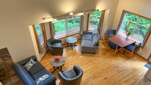 A spacious living area with a wooden floor, multiple seating arrangements including sofas and chairs, a round coffee table, and large windows offering a view of greenery. - The River House' w/ Deck, on McKenzie River! (Springfield)