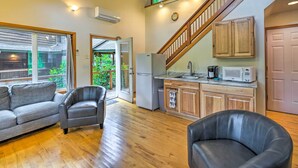 A spacious living area with a grey sofa, leather armchairs, wooden flooring, a kitchen with wooden cabinets, a white refrigerator, a microwave, and a staircase leading to an upper level. - The River House' w/ Deck, on McKenzie River! (Springfield)